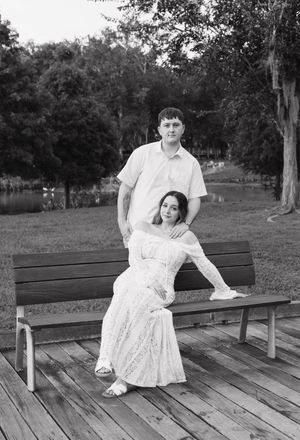 Black and white photo of a couple posing on a park bench, woman in lace dress seated, man standing behind her.
