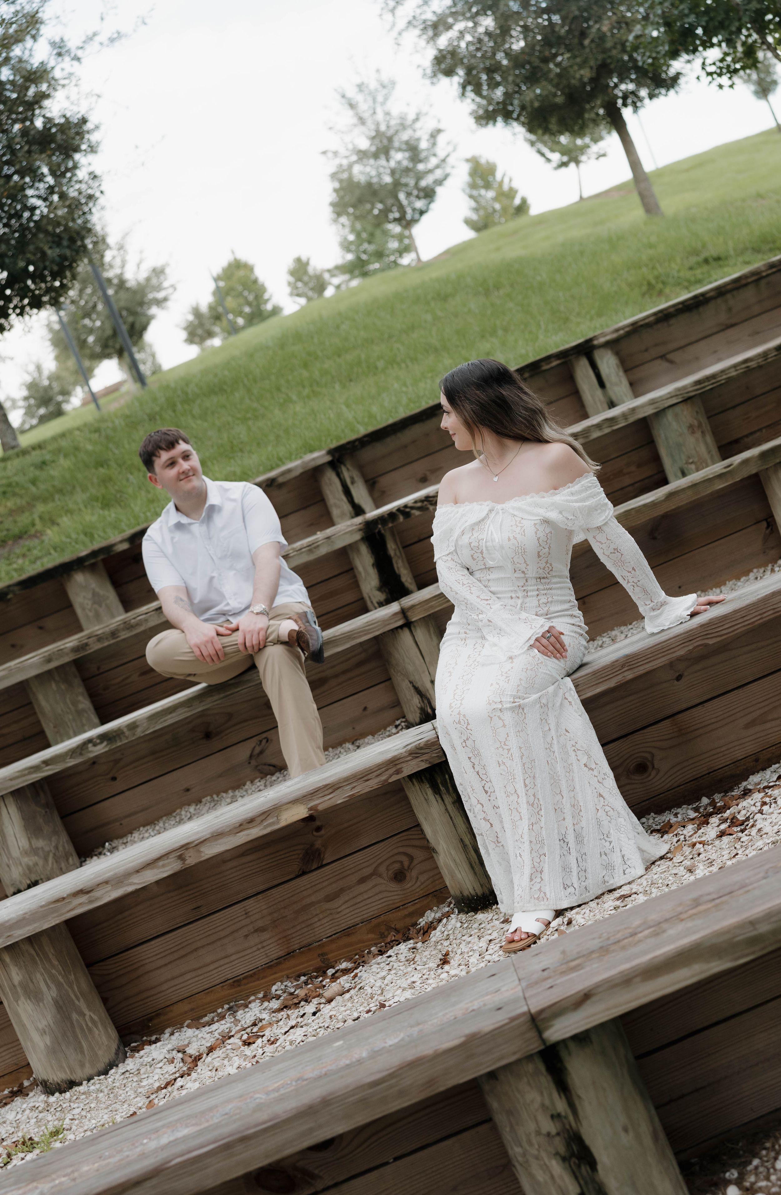 A couple in white attire poses on wooden bleachers outdoors, with a green grassy hill in the background.