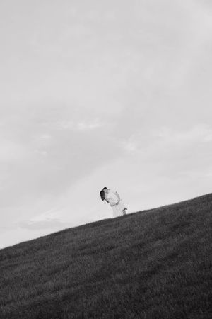 Black and white photo of a couple embracing on a grassy hillside under a cloudy sky.