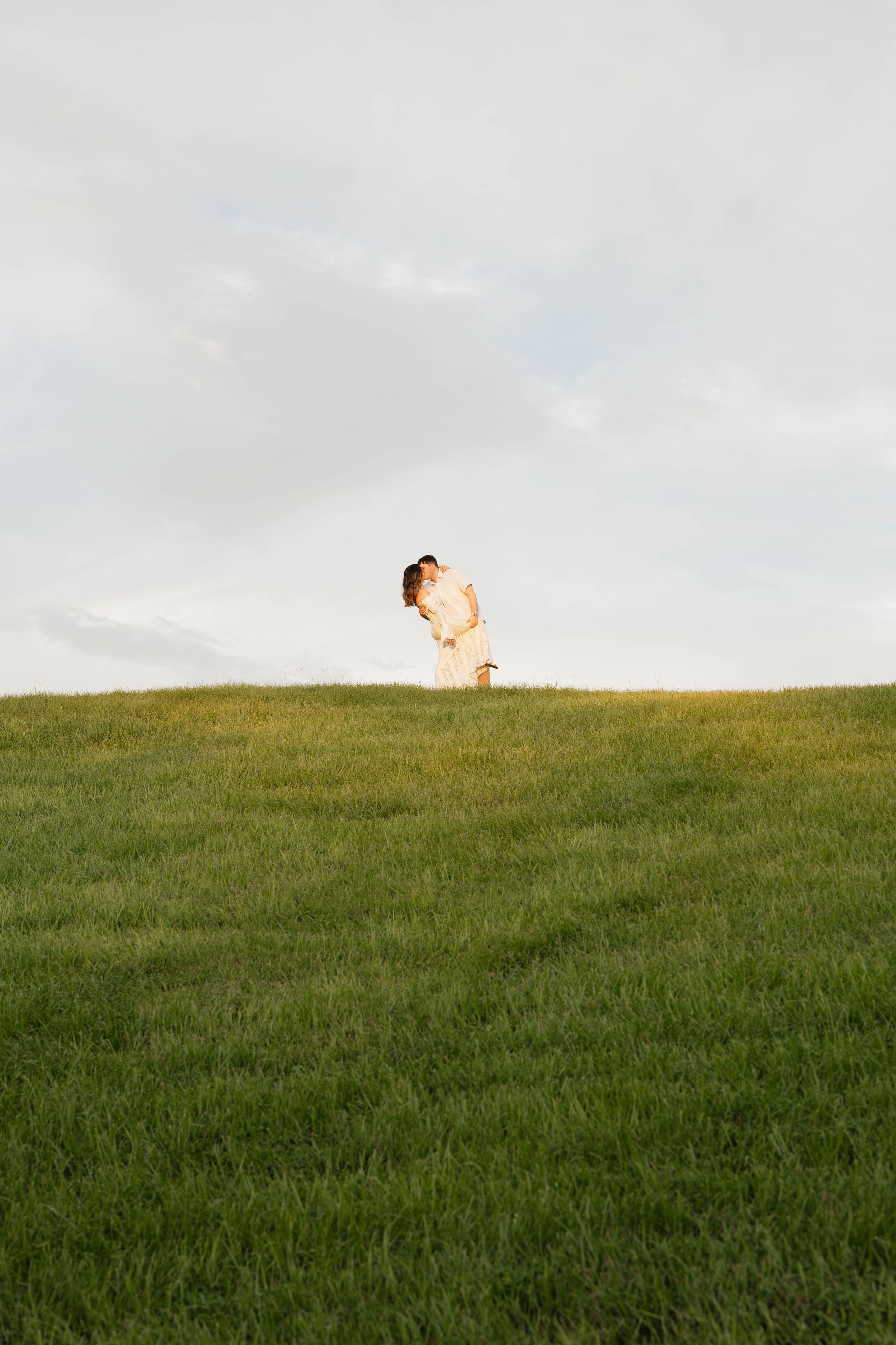 A couple embraces on a lush green hill under a soft cloudy sky during golden hour.
