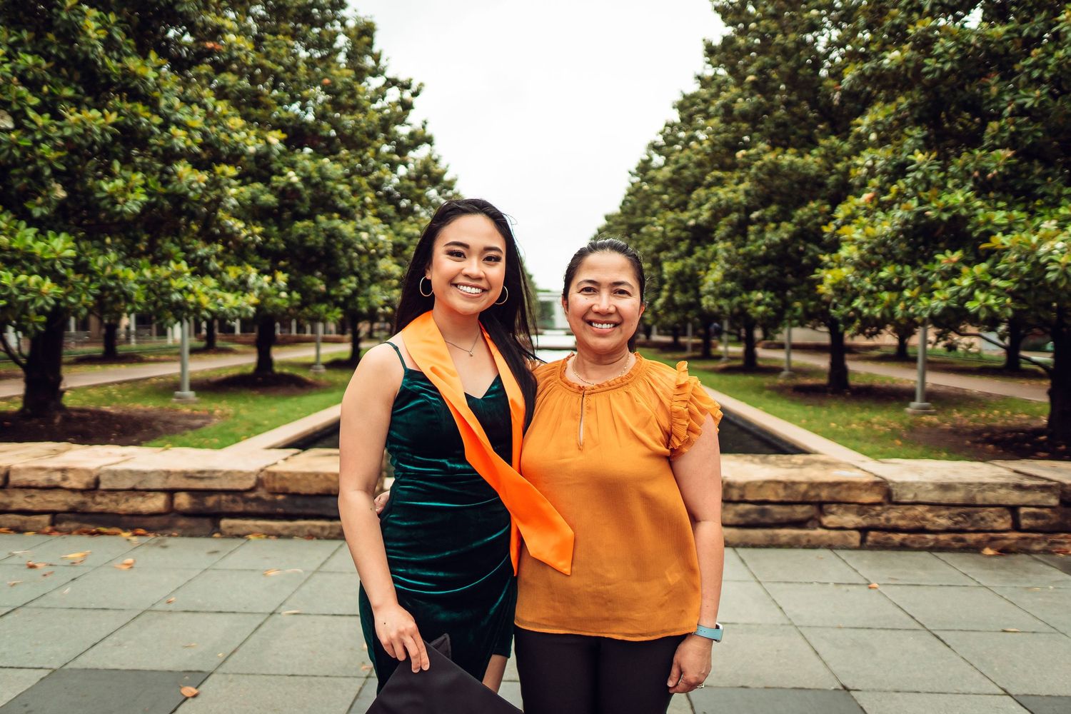 Two students in orange and green attire pose together on campus grounds with trees in background. UTD Graduate Portraits by Eric Aaberg Phot