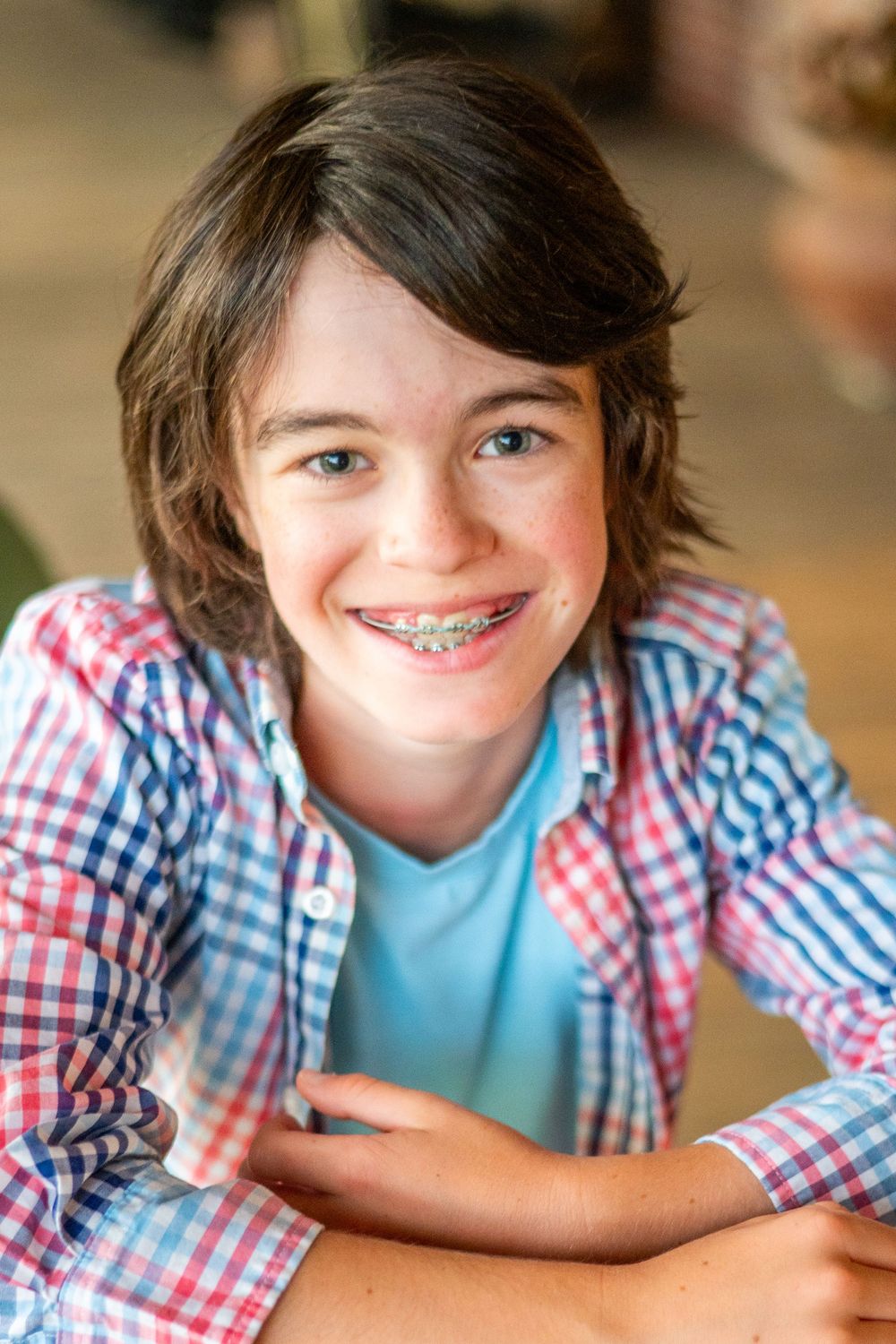 Young person wearing braces smiles warmly in a blue shirt and red plaid button-up while sitting at a table.