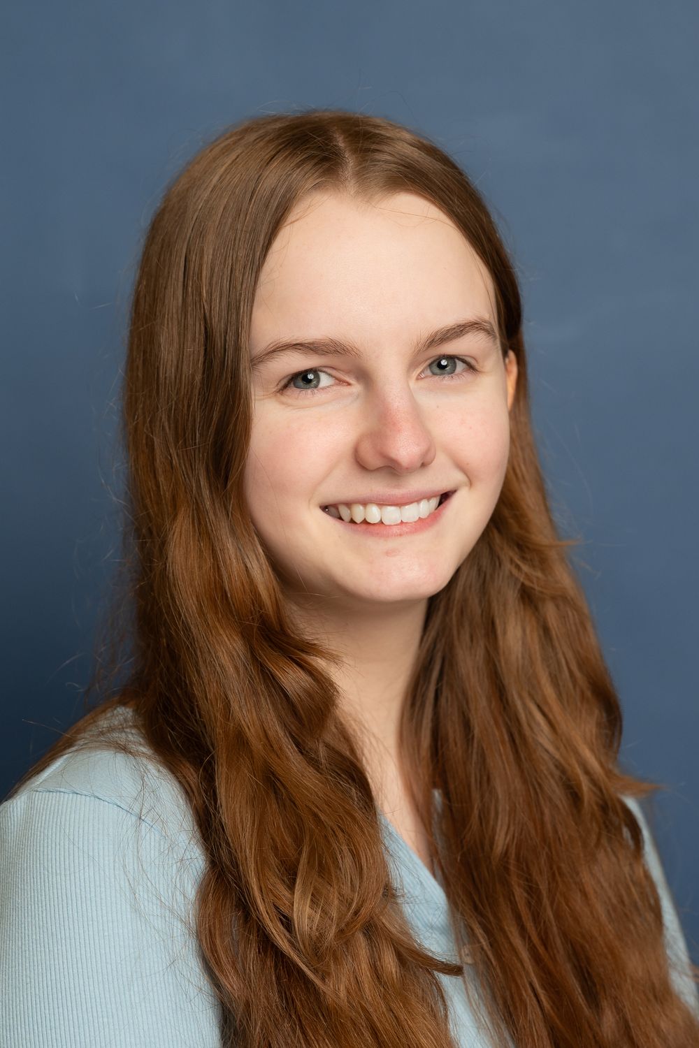 A person with long brown wavy hair wearing a light blue sweater smiles against a blue background.