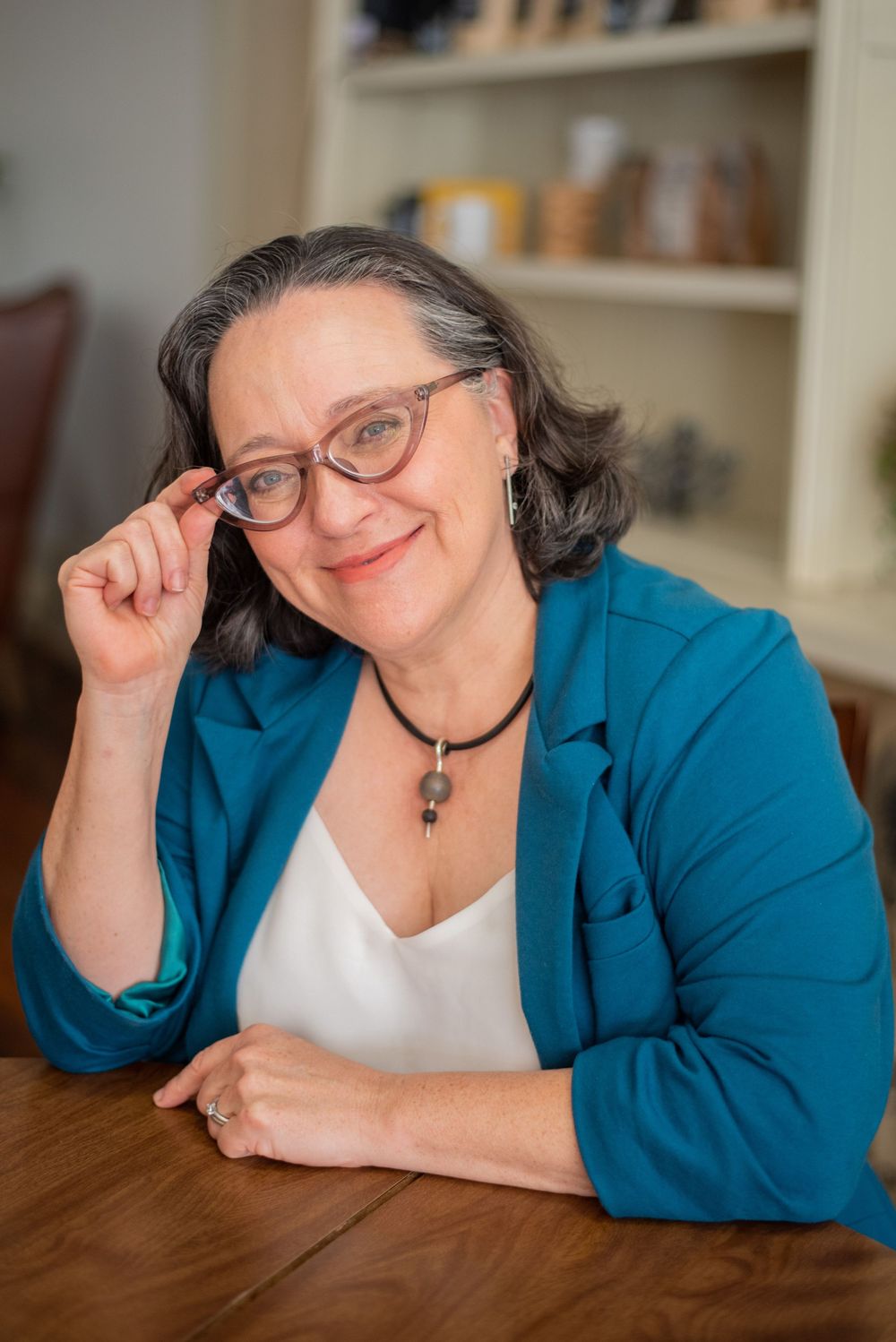 A person wearing a bright blue blazer and glasses poses playfully while leaning on a wooden table.