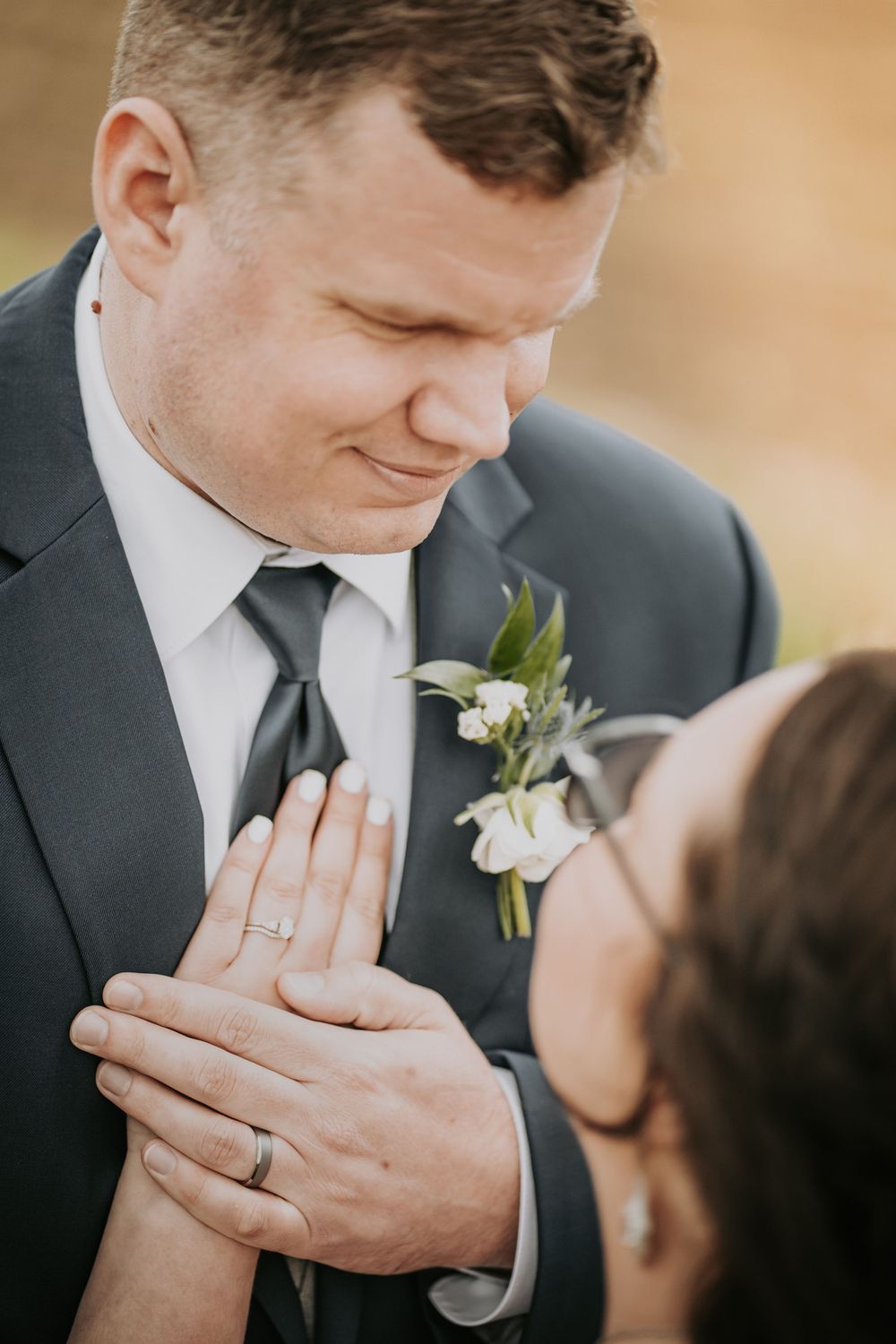 Intimate wedding moment showing clasped hands with matching rings and a white boutonniere on dark suit.