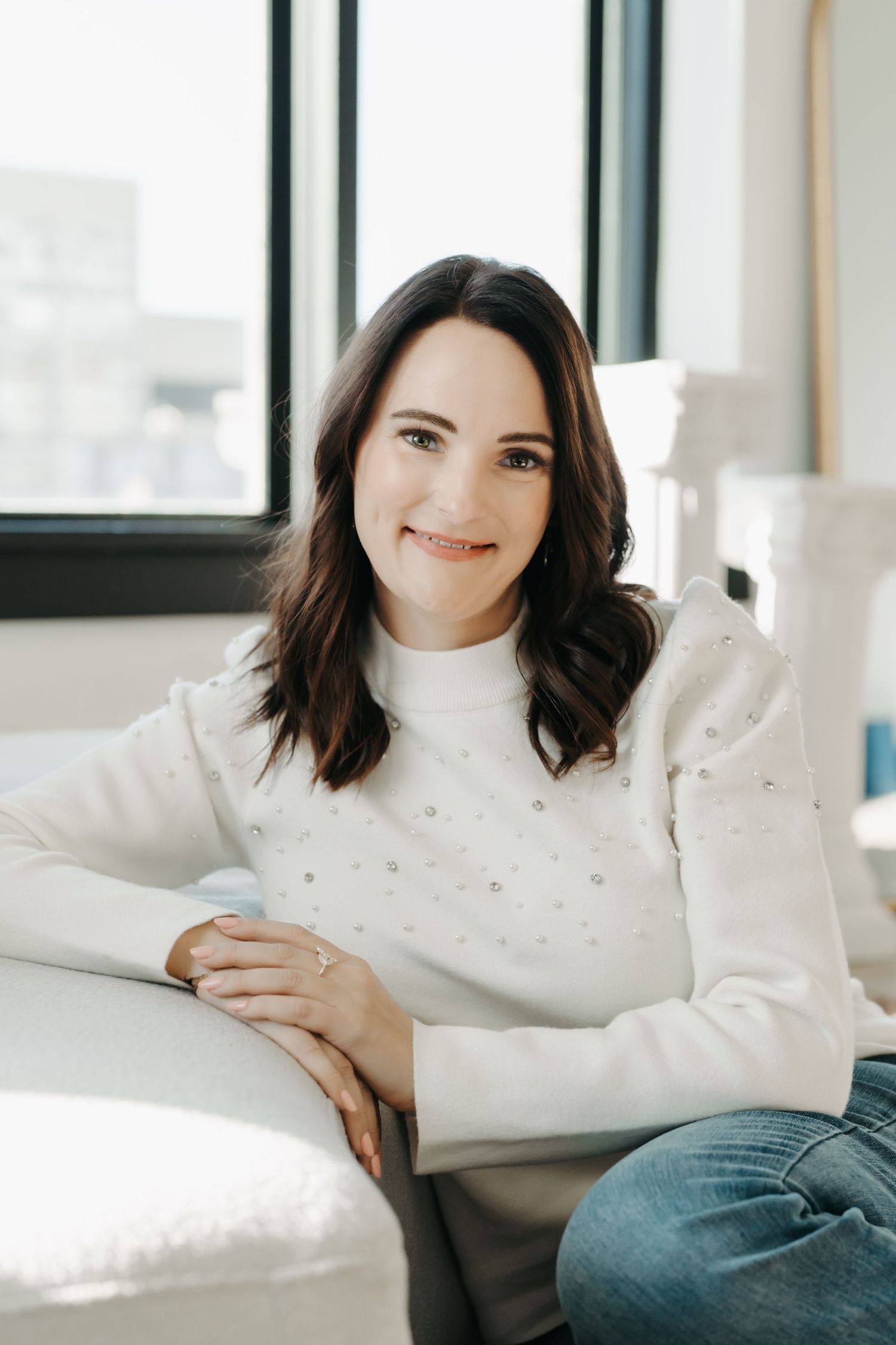 A smiling brunette woman in a white pearl sweater and jeans sits relaxed on a light sofa near a bright window.