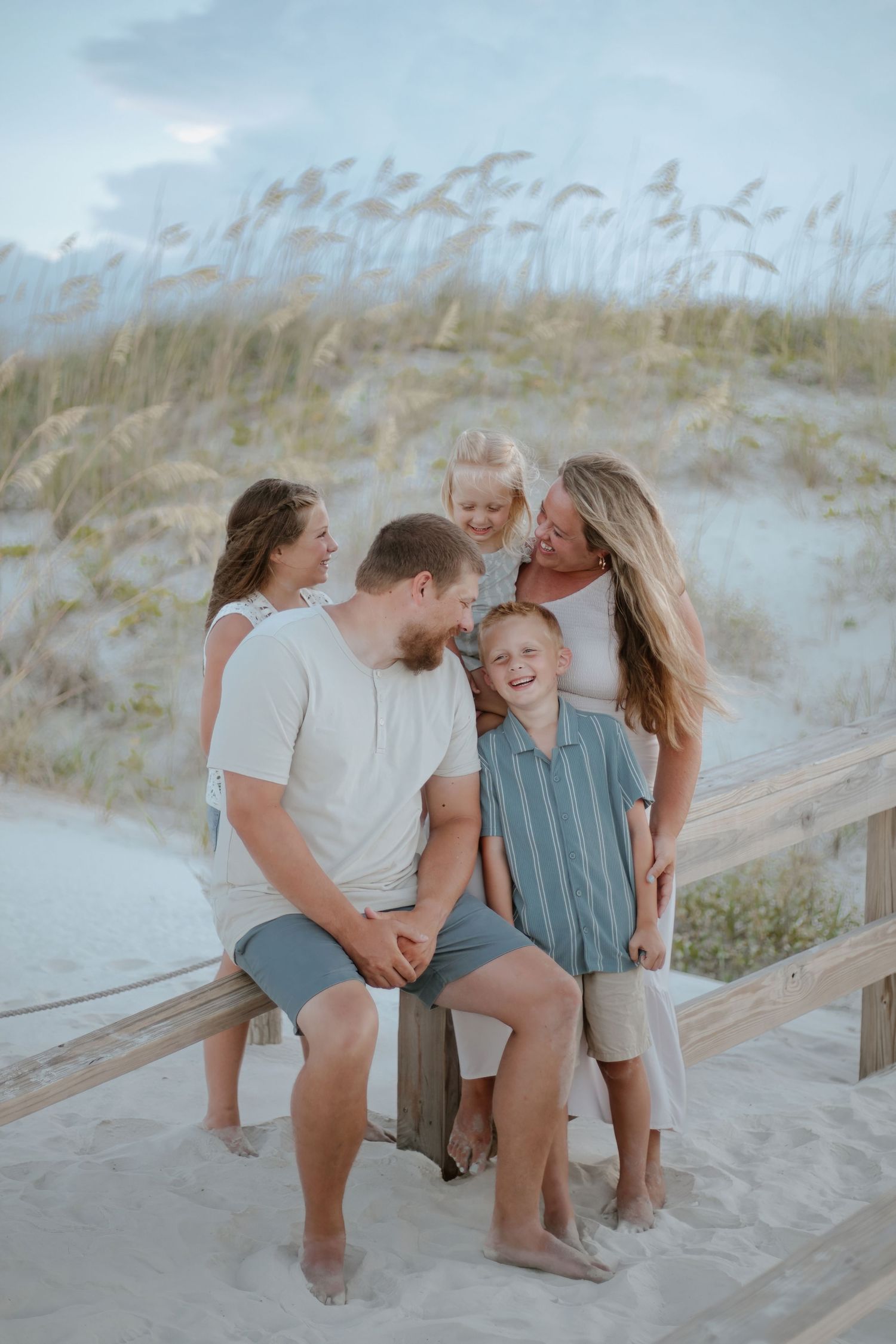 Playful family photos on wooden boardwalk at beach with sand dunes and sea grass in background.