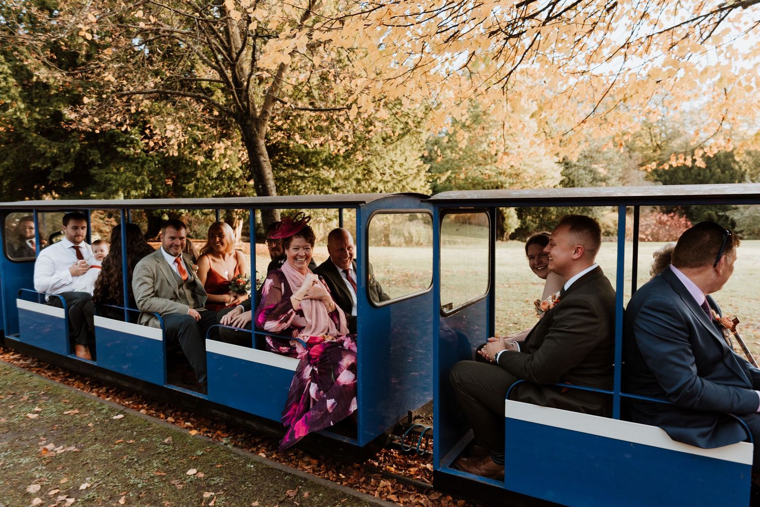 Wedding guests ride in blue and white train cars through an autumn park setting during a celebration.