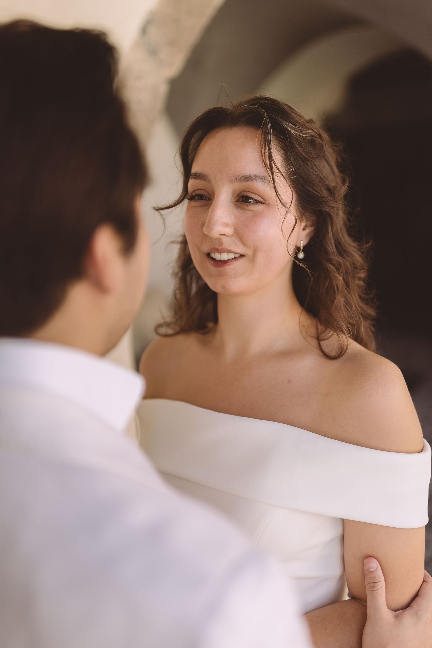 A bride in an off-shoulder white dress smiles warmly at her groom at their Santorini wedding.