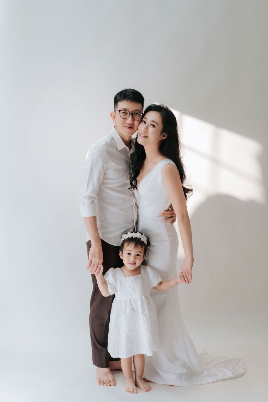 Asian family portrait with man, pregnant woman in white dress, and toddler girl in white studio setting.亞洲家庭合影，照片中有一位男士、一位身穿白色連身裙的孕婦和一位小女孩，拍