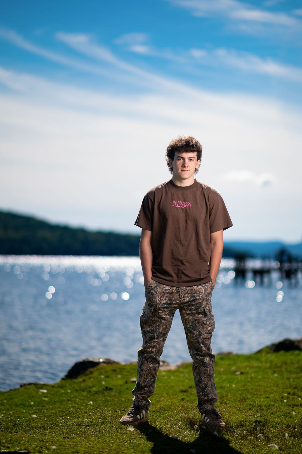 Person in brown shirt and camo pants stands on grassy hill overlooking water on sunny day.
