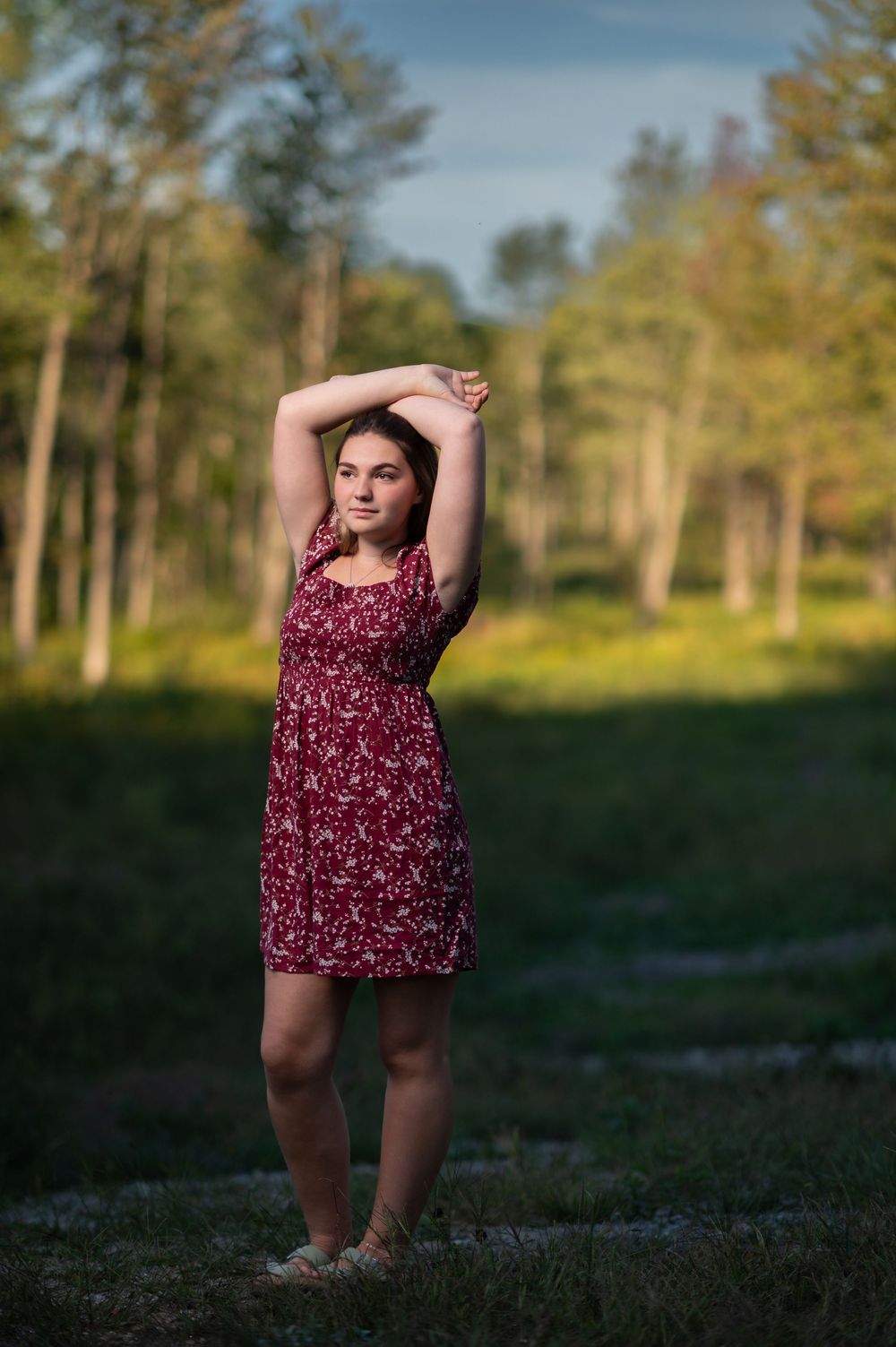 Figure in burgundy dress poses against natural landscape at sunset.