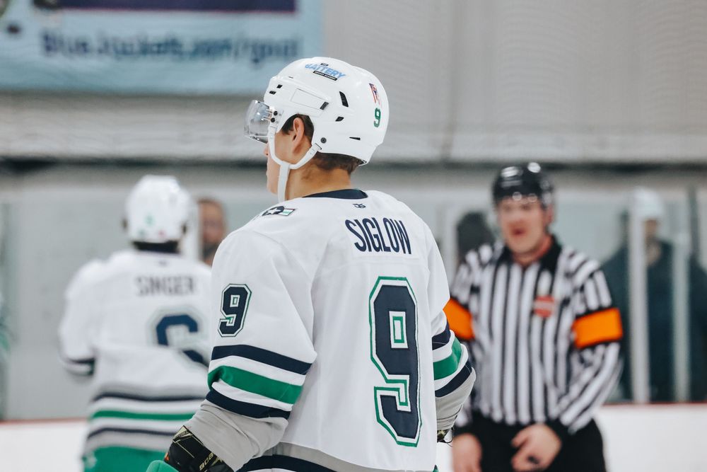 Hockey players in white and dark uniforms chase after the puck on an indoor rink.