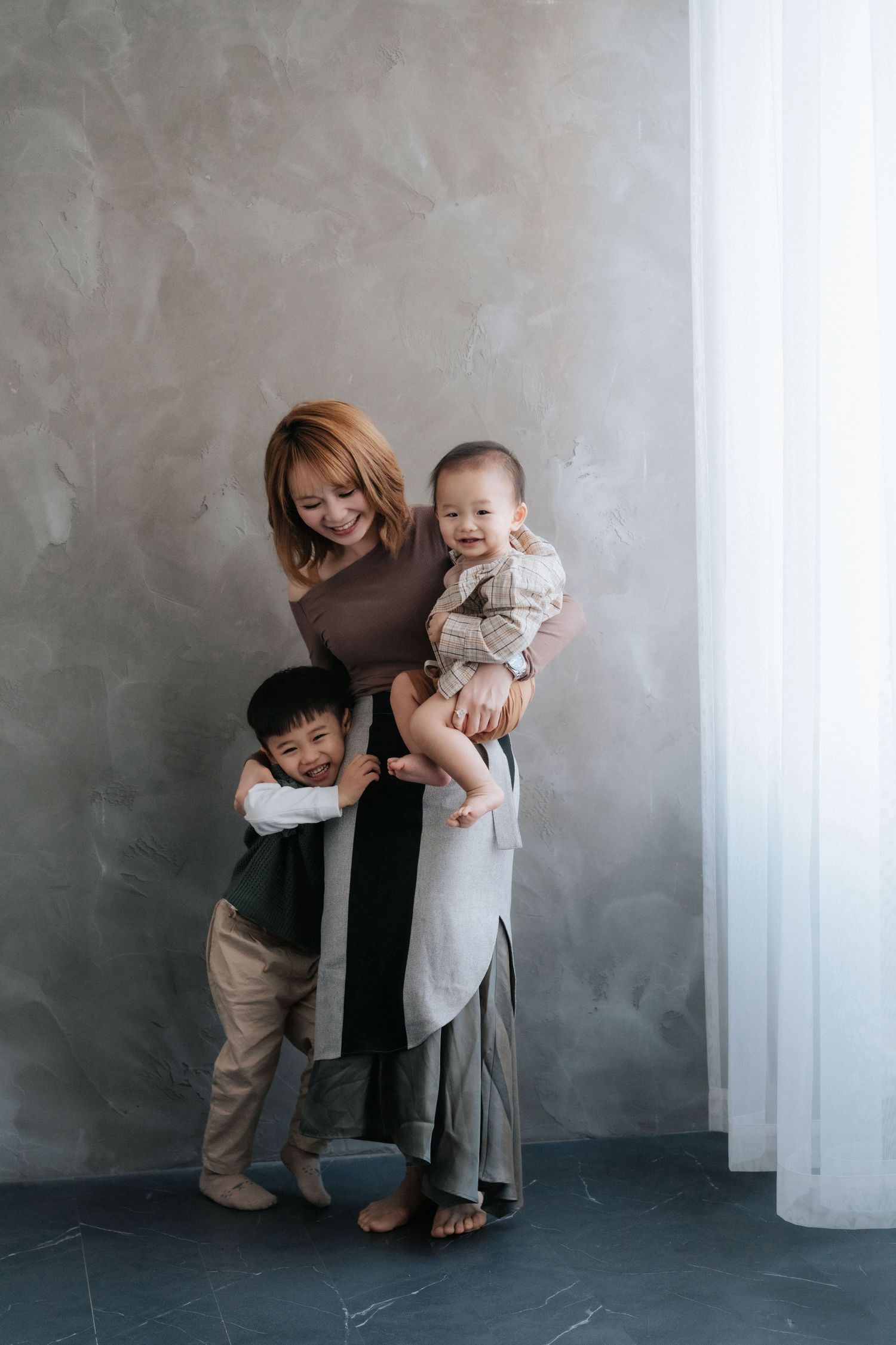A smiling Asian mother holds a baby while her young son hugs her legs, standing by a gray textured wall.一位微笑的亞洲母親抱著嬰兒，她的小兒子抱著她的腿，站在一面灰色紋理的牆邊