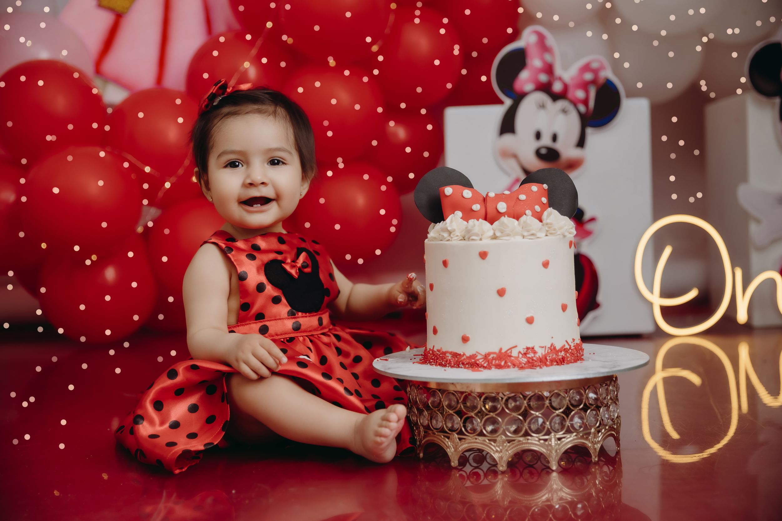 Minnie Mouse themed first birthday party setup with white cake, red balloons, and polka dot decorations against red backdrop.