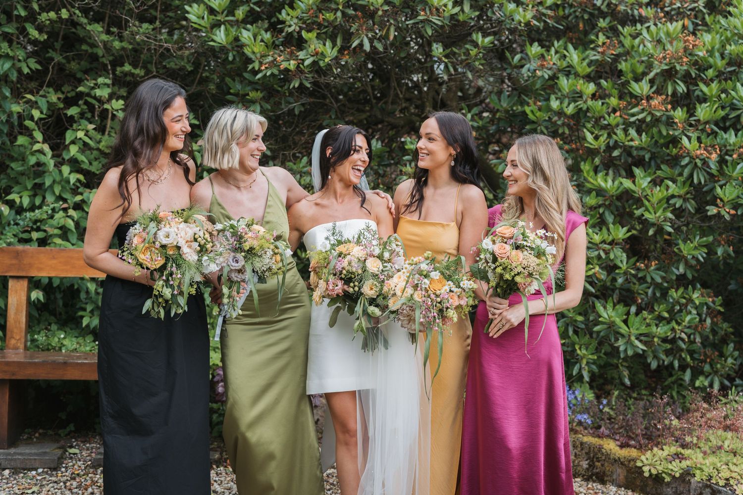 Bridesmaids in olive green and pink dresses gather around bride in white gown during outdoor garden wedding celebration.