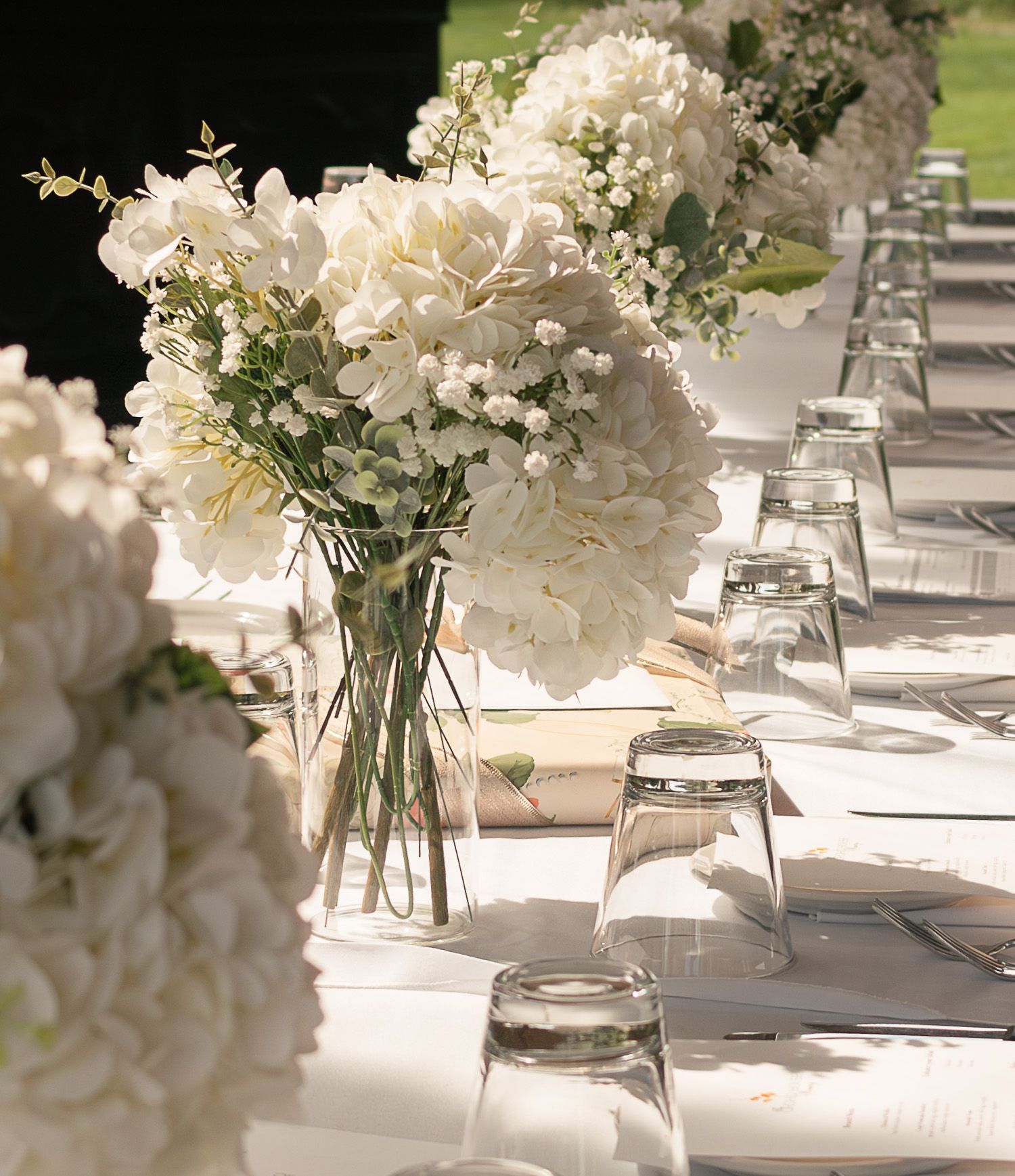 Elegant white hydrangea centerpieces in glass vases line a wedding reception table with white tablecloths.