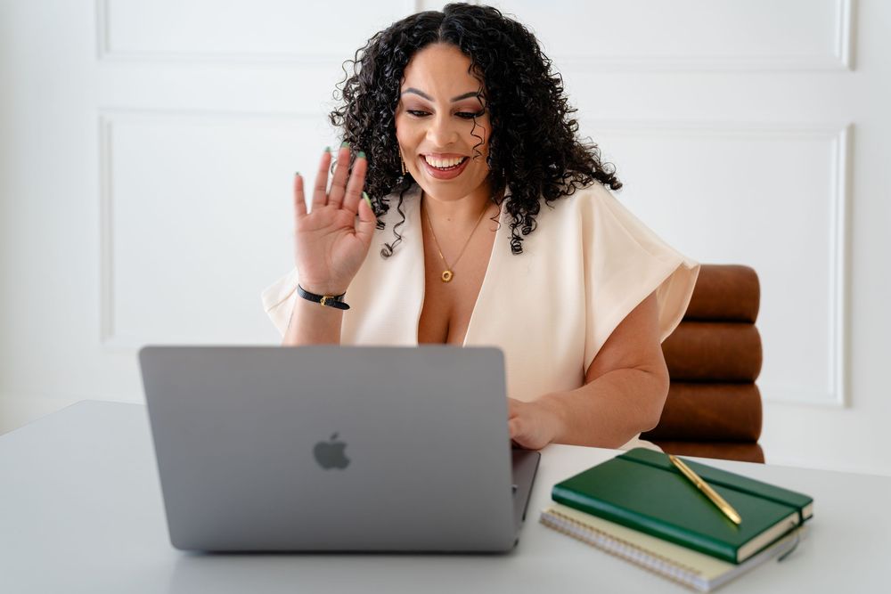Professional working remotely waves during video conference call while using laptop computer at desk.