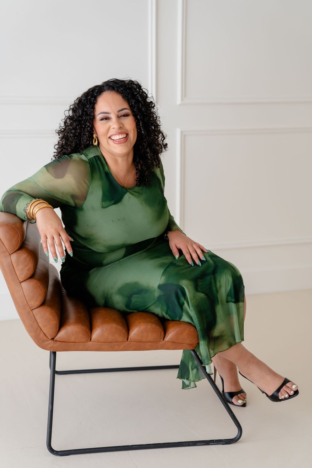 Elegant emerald green dress worn while relaxing on a tan leather modern chair against white wall.