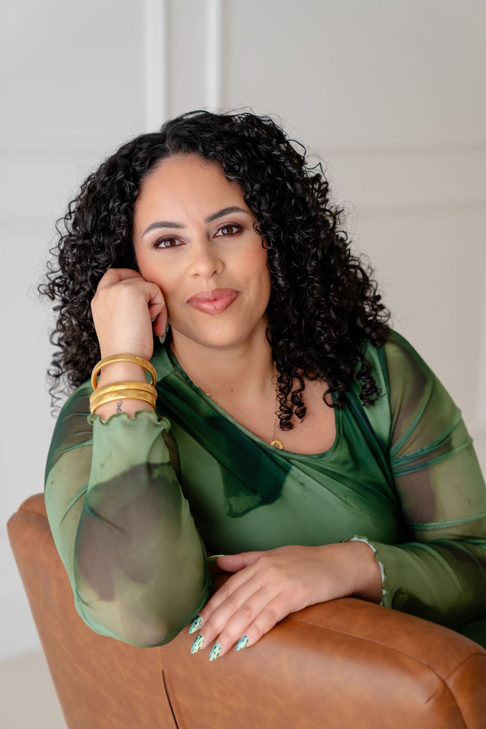 Portrait of someone with curly dark hair wearing a green sheer top and gold bangles, sitting on brown leather furniture.