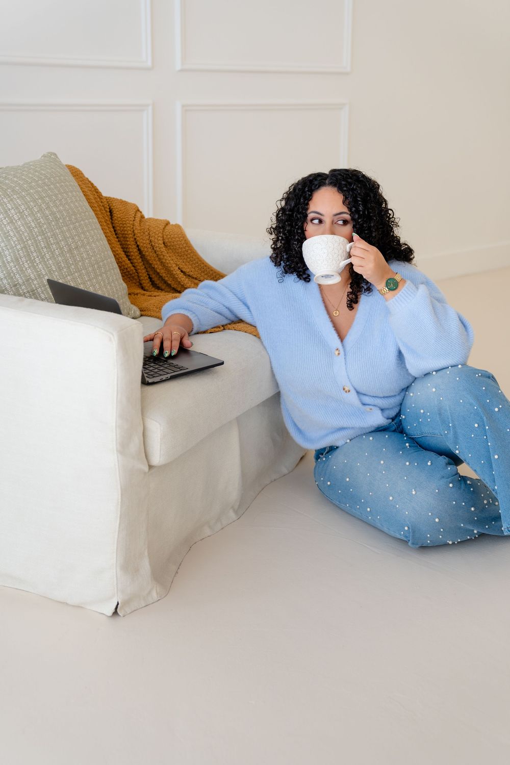 Person relaxing on white couch wearing blue cardigan and polka dot pants while drinking from white mug.