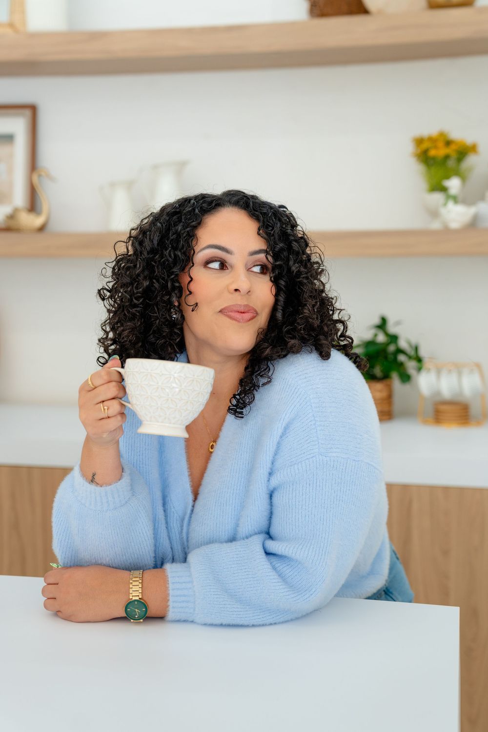Person in light blue sweater relaxing at white counter while drinking from teacup in bright modern kitchen setting.