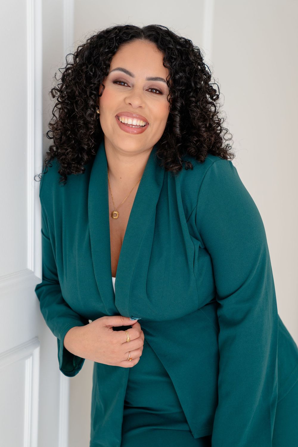 Professional portrait of someone wearing a teal blazer and curly hair posing against a white wall.