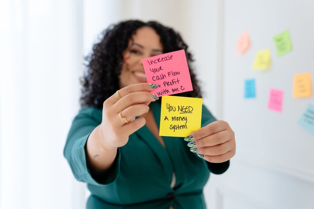 Person in green sweater holds colorful sticky notes against wall with more notes in background.