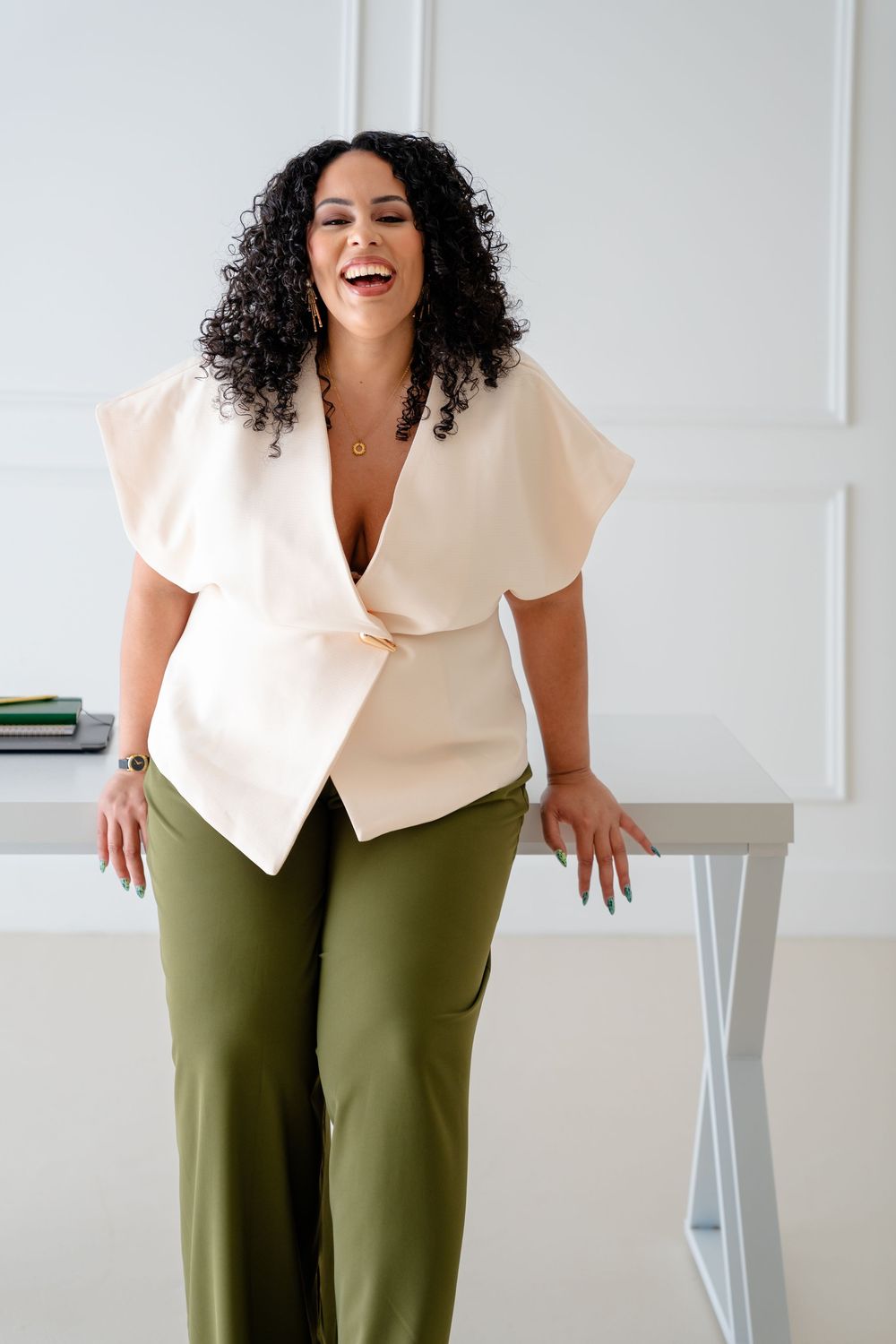 A curly-haired person in a cream wrap top and olive pants stands laughing joyfully against a white wall.