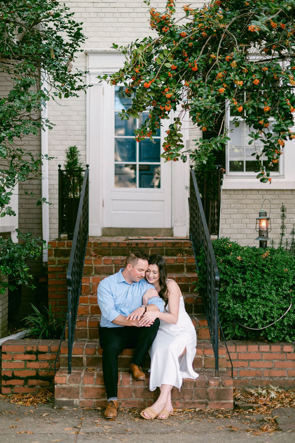 Romantic couple sitting together on a brick ledge beneath orange flowering trees and white building exterior.