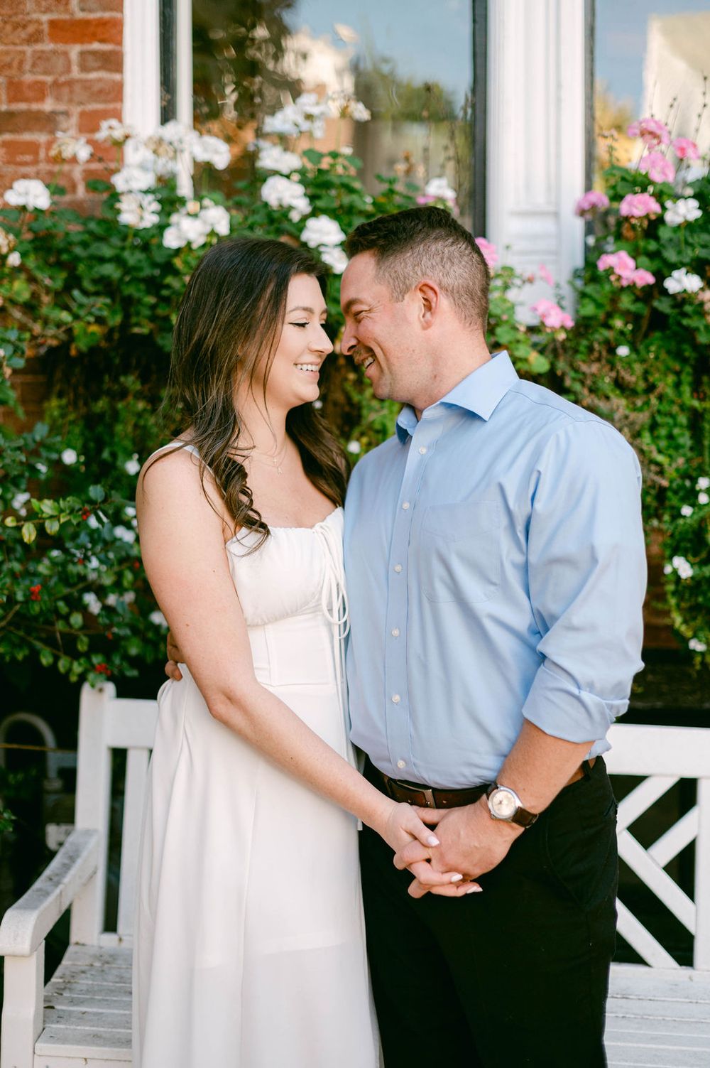 A romantic couple shares tender moments in front of a white porch with pink flowers and brick walls.