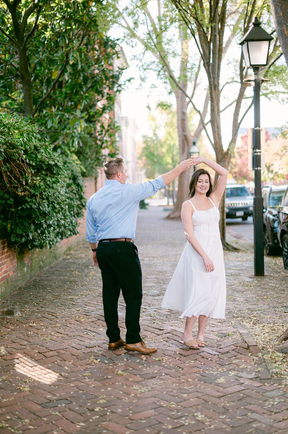 A couple walks along a charming brick sidewalk lined with trees and lamp posts in a romantic outdoor setting.