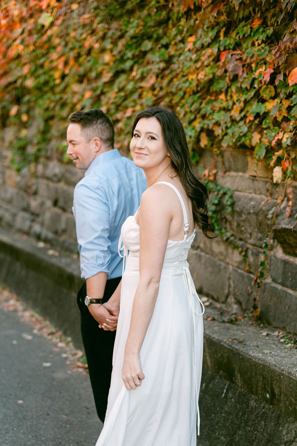A couple in white and blue attire poses against a rustic stone wall with autumn foliage.