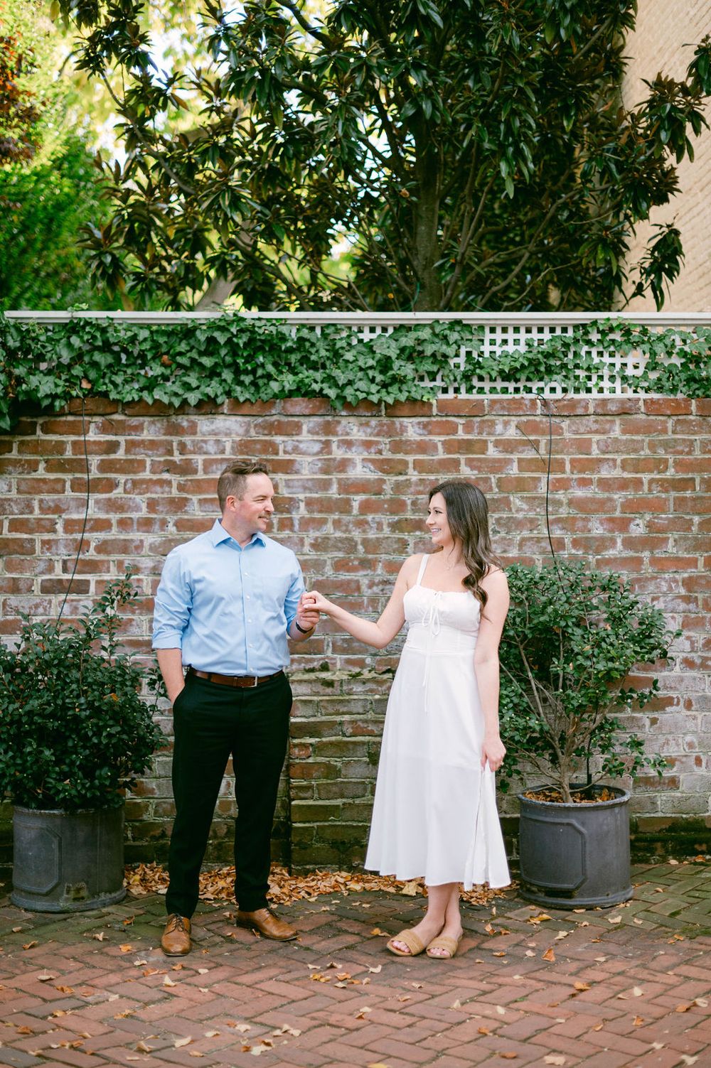 Couple in white dress and blue shirt posing playfully against a brick wall with greenery.