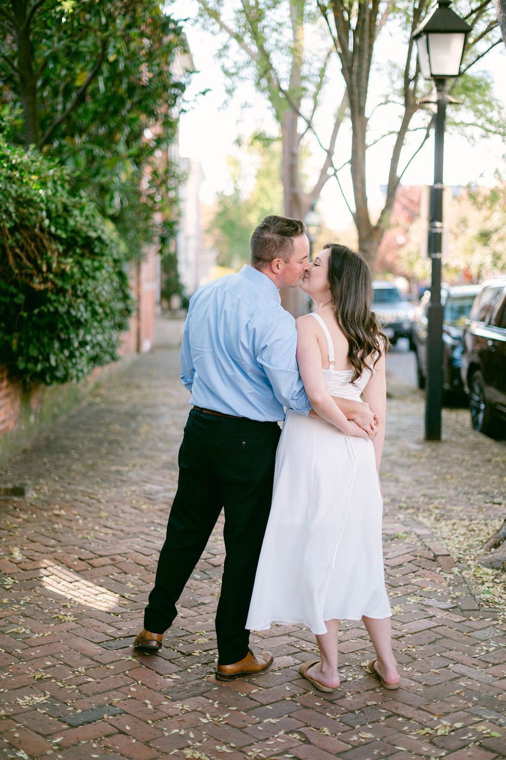 A romantic couple shares intimate moments on a cobblestone path lined with trees and lamp posts.