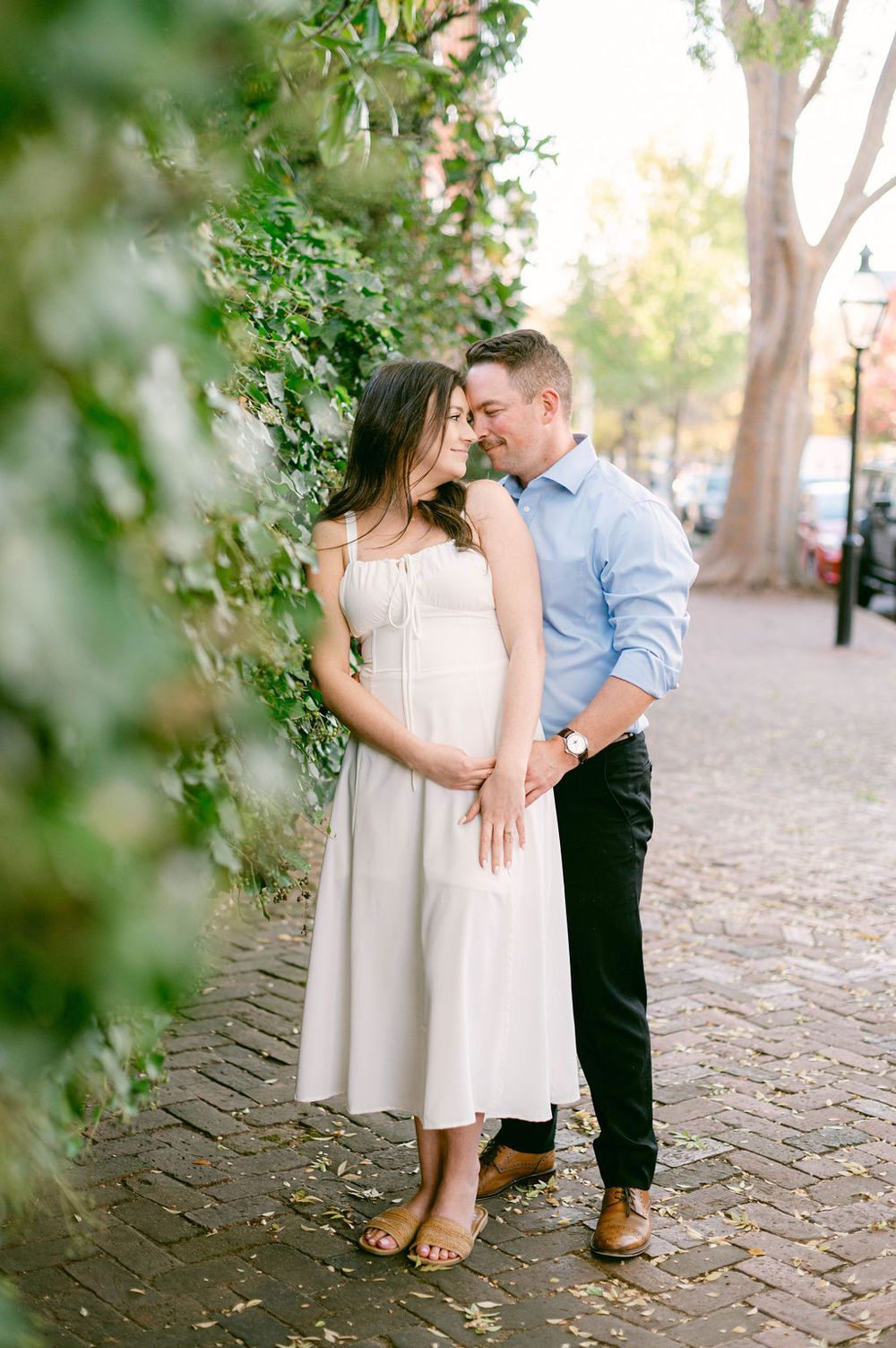 A couple embraces tenderly on a brick walkway surrounded by lush greenery and dappled sunlight.