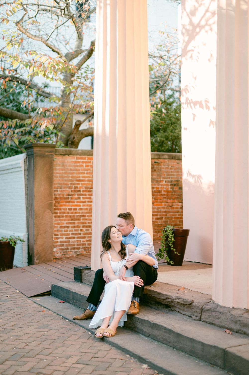 Romantic couple sitting and laughing together on historic building steps with brick walls and white columns.