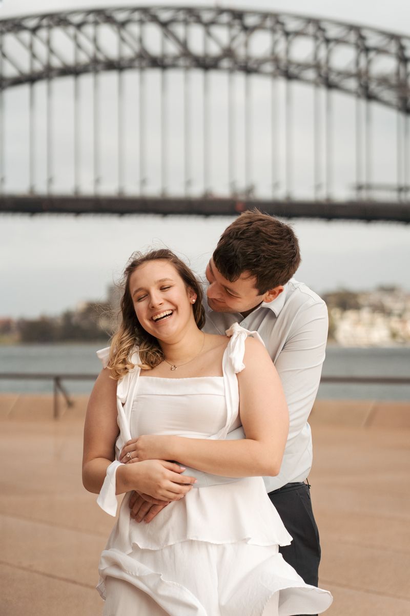 A couple shares intimate moments and laughter together with the harbour bridge in the background during their engagement photoshoot