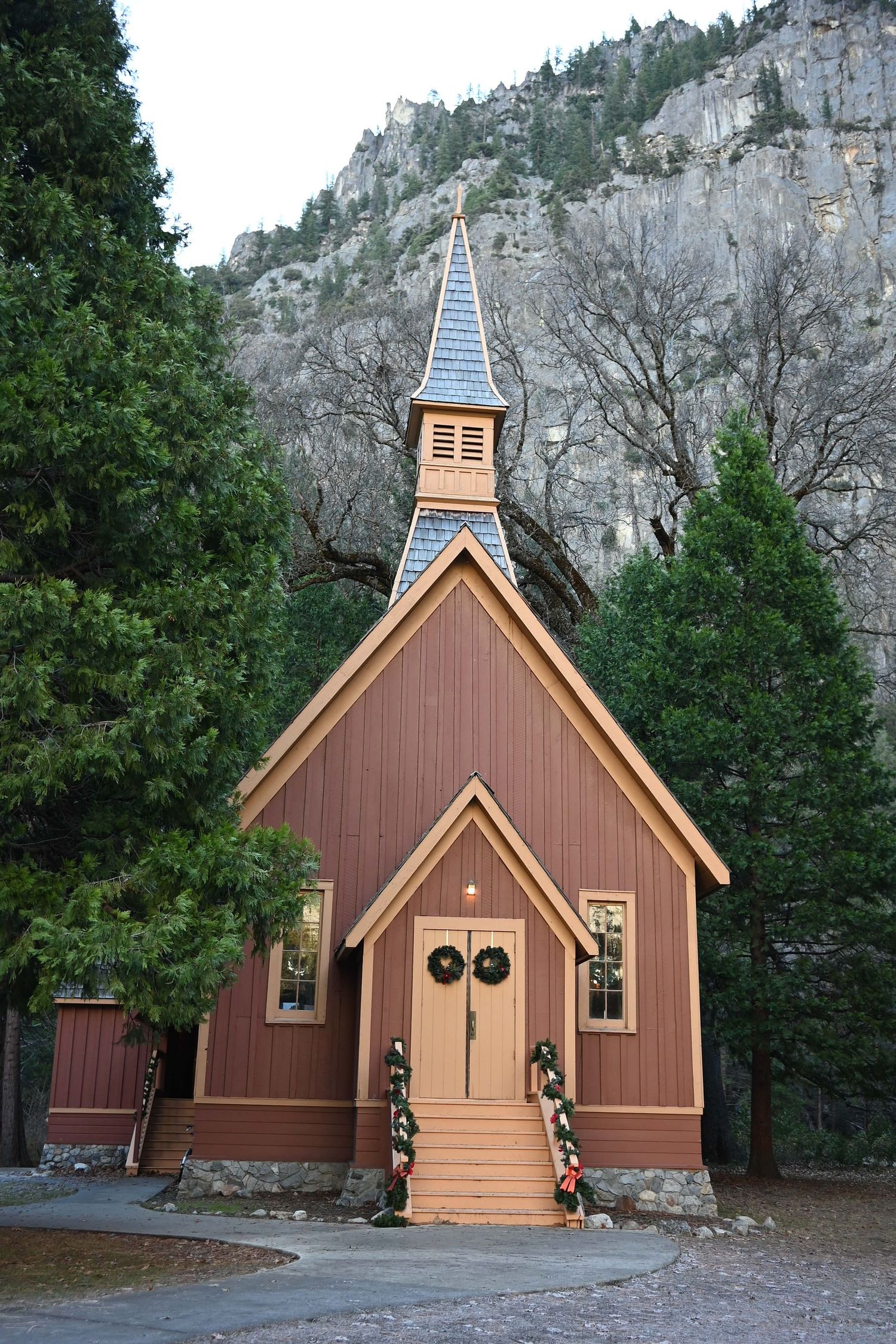 Historic wooden chapel with brown exterior and blue steeple sits nestled against towering granite cliffs in Yosemite Valley.