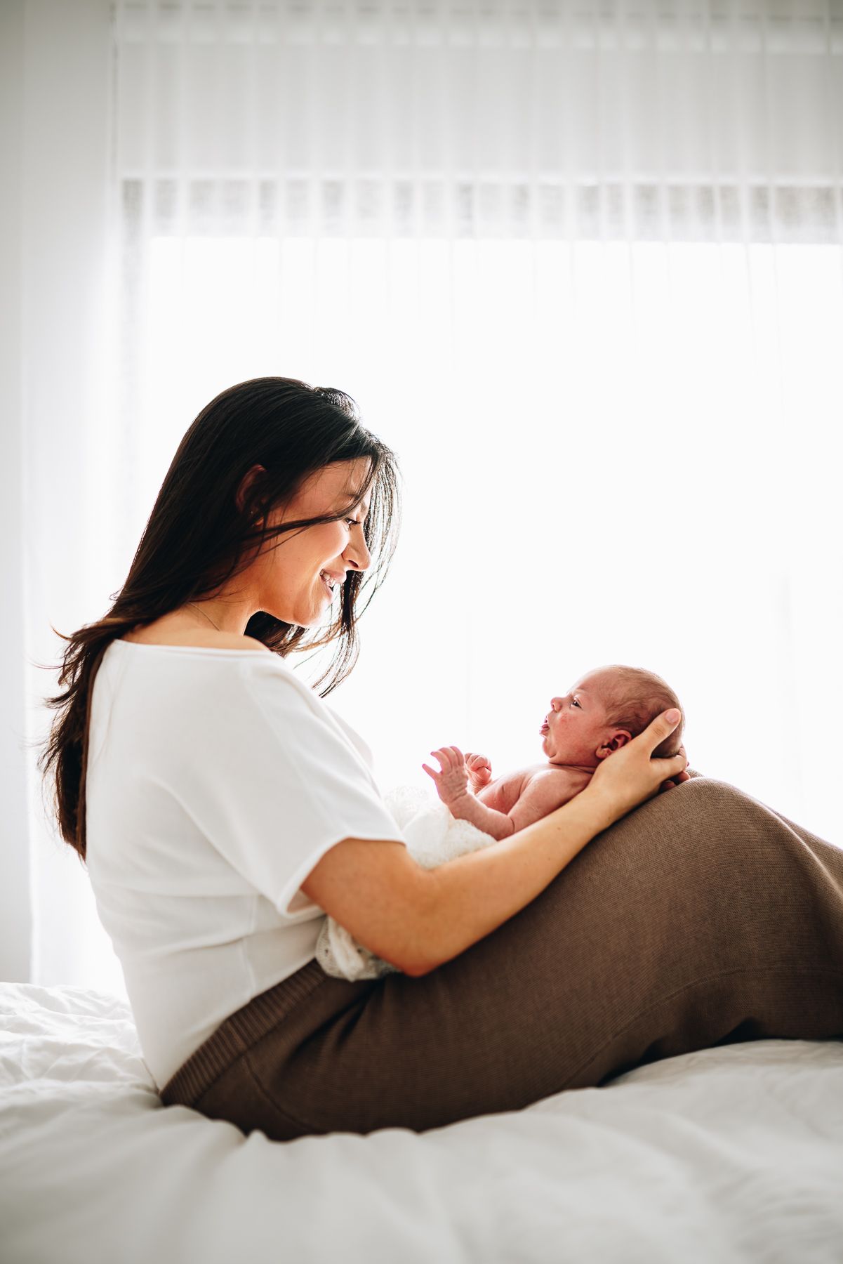 A first time mom sitting on a bed cradling and playing with her newborn, taken by an in-home newborn photographer in Melbourne.