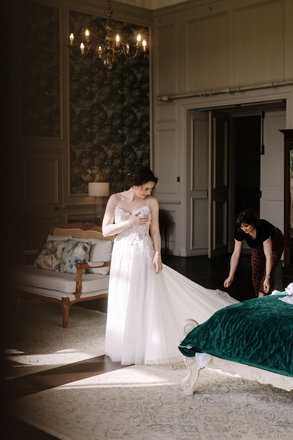 A wedding dress preparation scene in an elegant room with chandelier and vintage furniture as sunlight streams through.