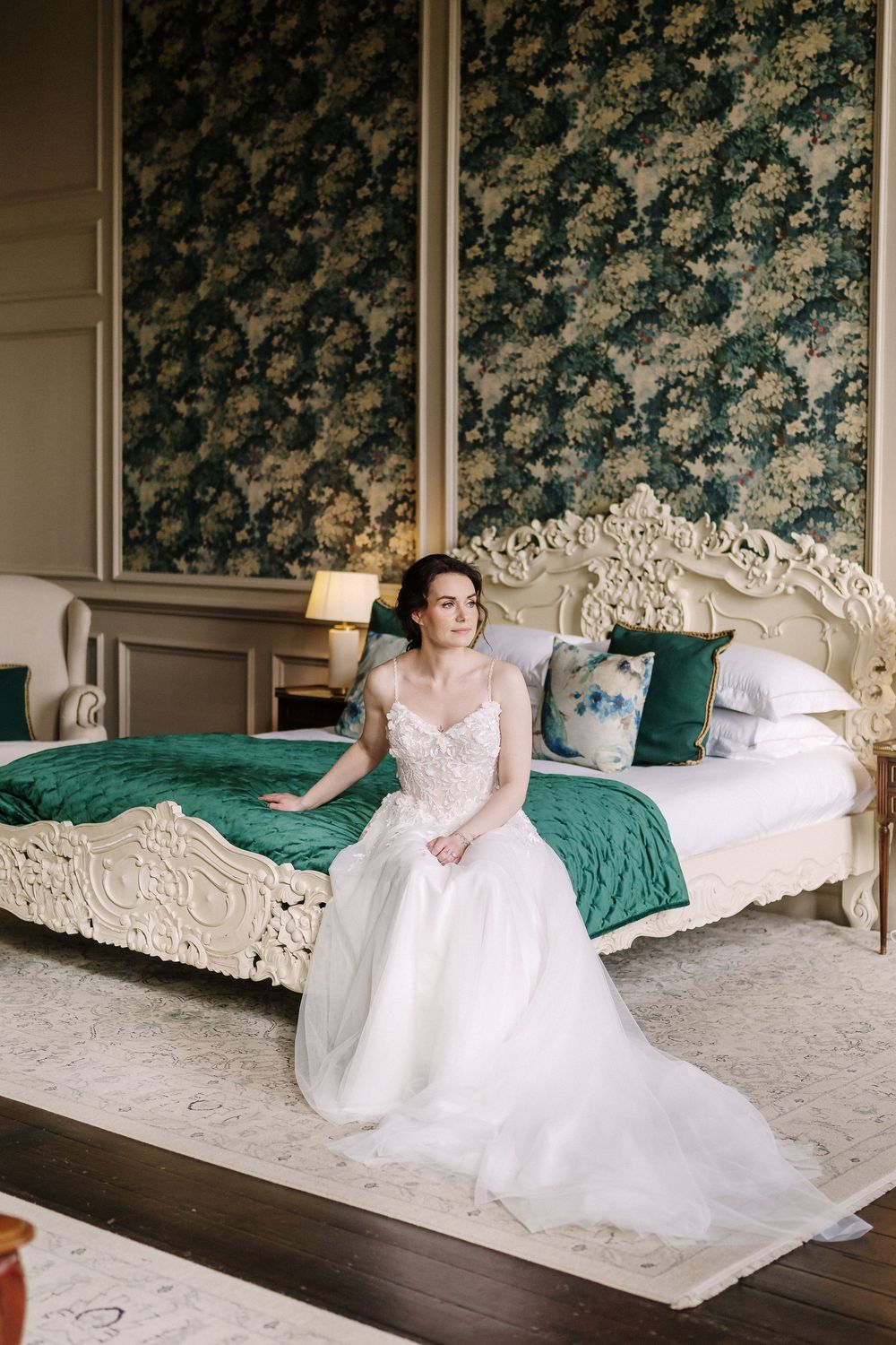 Elegant white lace wedding gown with illusion sleeves displayed on ornate vintage bed with emerald green bedding.