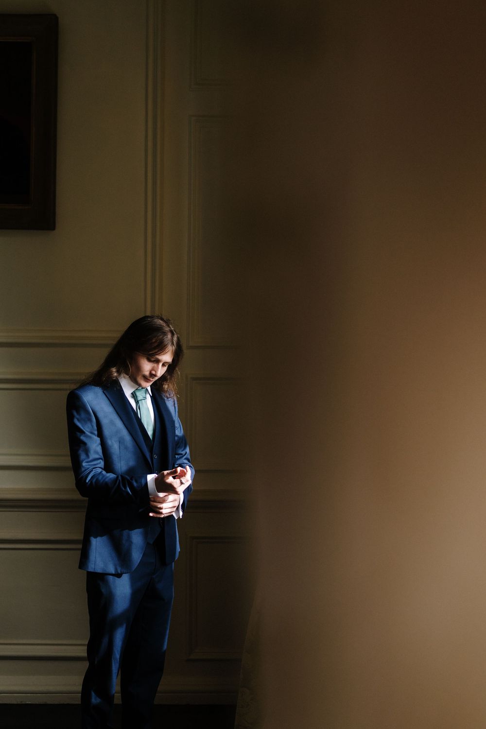 Person in a navy blue suit stands in dramatic shadow against a beige wall while looking at a small object.