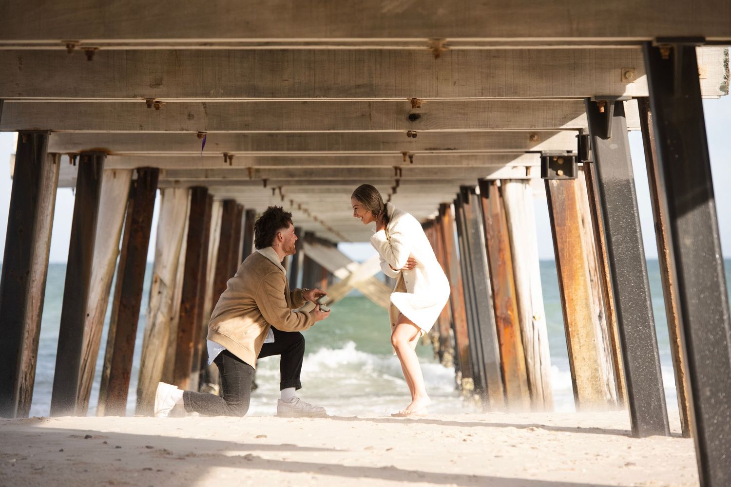 Man proposing to woman on sandy beach under a pier with ocean waves in the background.