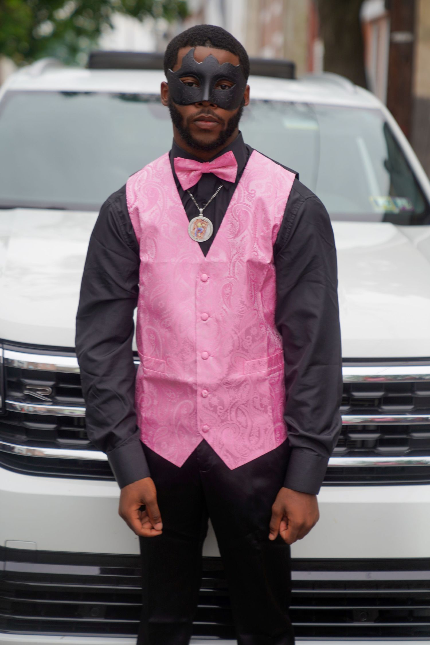 A person wearing a black shirt and pink vest poses in front of a parked car.