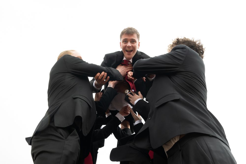 A group of people in black suits huddle together in a playful low-angle shot against a white sky.