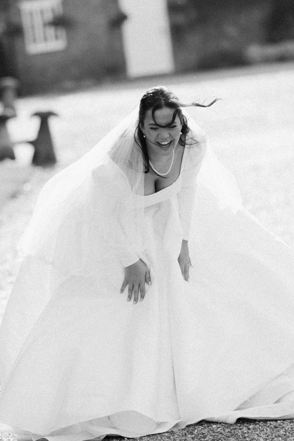 A joyful bride in a flowing white wedding dress and veil laughs as her dress billows in the wind.