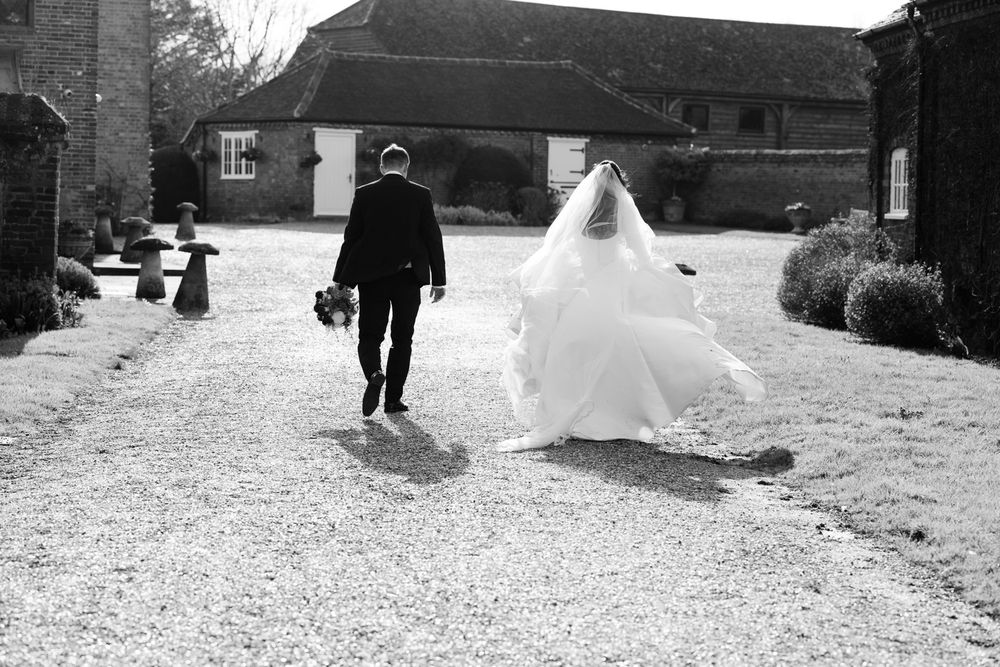 A black and white wedding photo showing a couple walking on a gravel path outside a rustic building.