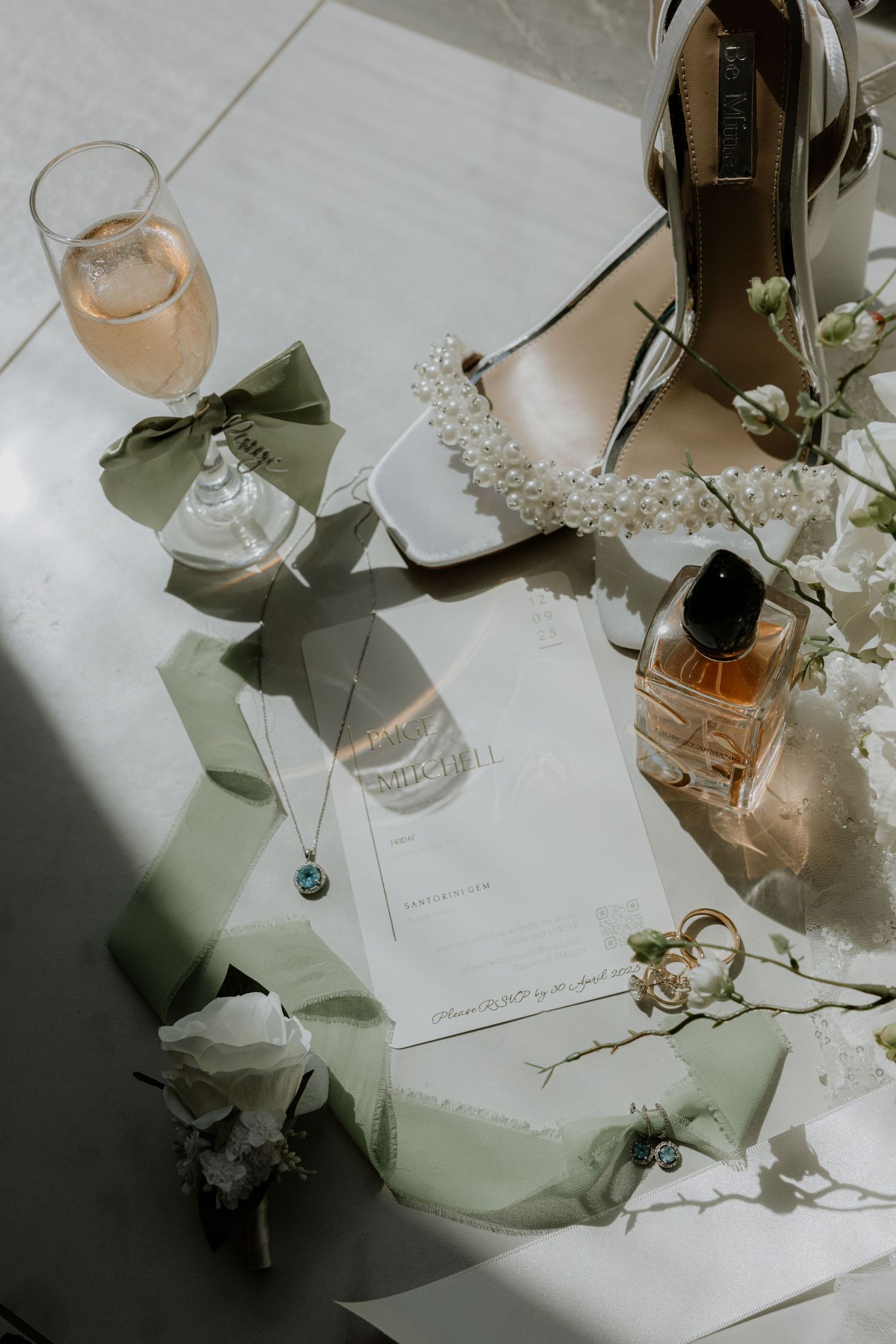 Elegant wedding details with champagne, perfume bottle, heels with pearls, and sage green ribbons on white surface.