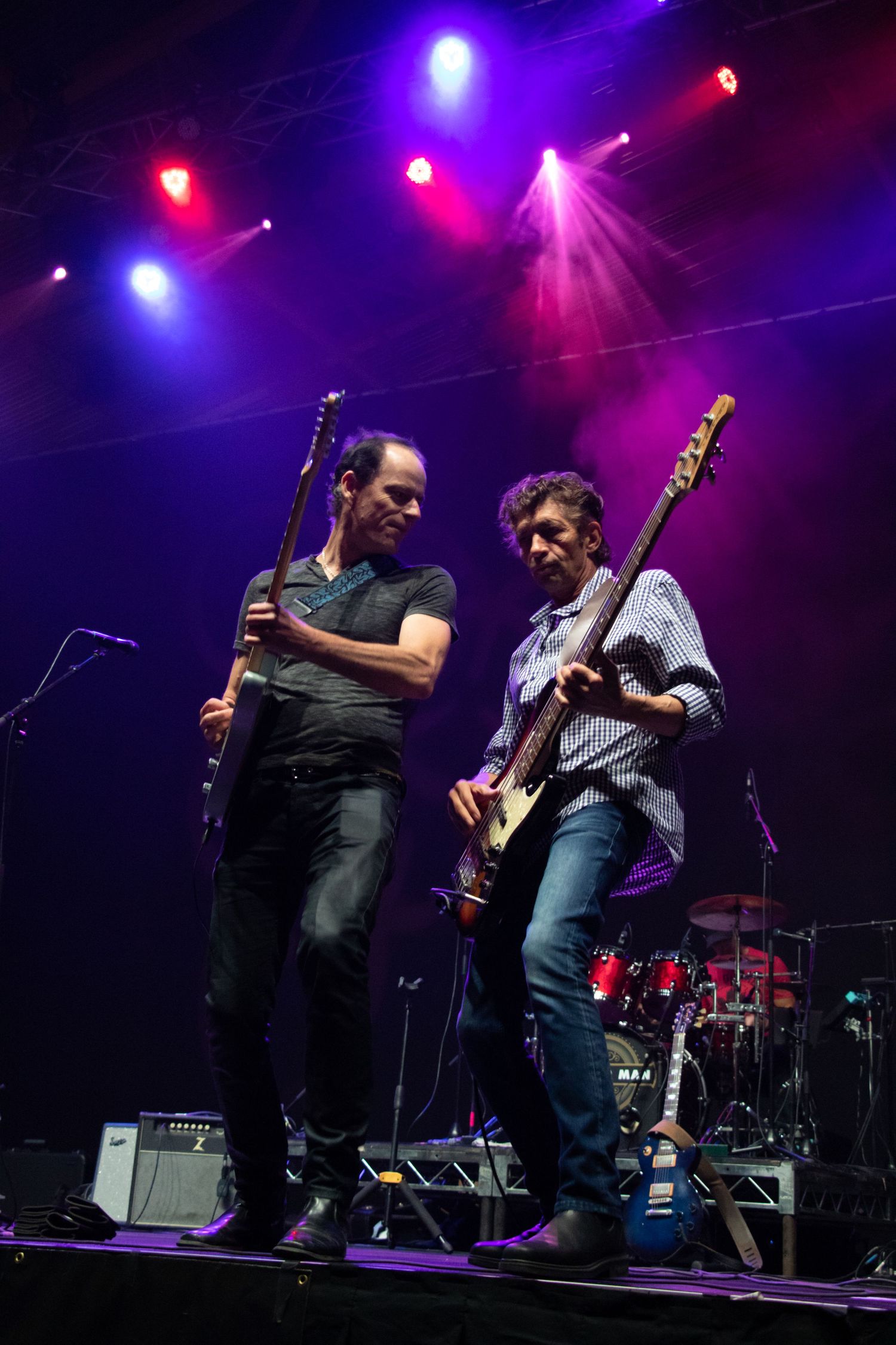 Two guitarists perform together on stage under dramatic purple and red concert lighting.